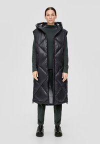 Schwarze gepolsterte Weste mit Kapuze, mit Rautensteppung und Frontreißverschluss. Getragen über einem dunkelgrünen Pullover und strukturierten Hosen, kombiniert mit Stiefeln.