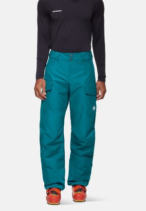 Personne portant un pantalon cargo teal avec poches à rabat, une chemise noire à manches longues et des bottes de ski orange vif, se tenant sur un fond blanc.