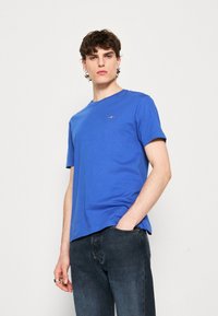 Blaues Baumwoll-T-Shirt mit kurzen Ärmeln und Rundhalsausschnitt. Verfügt über ein kleines Logo im Bereich der linken Brust. Kombiniert mit dunklen Jeans.