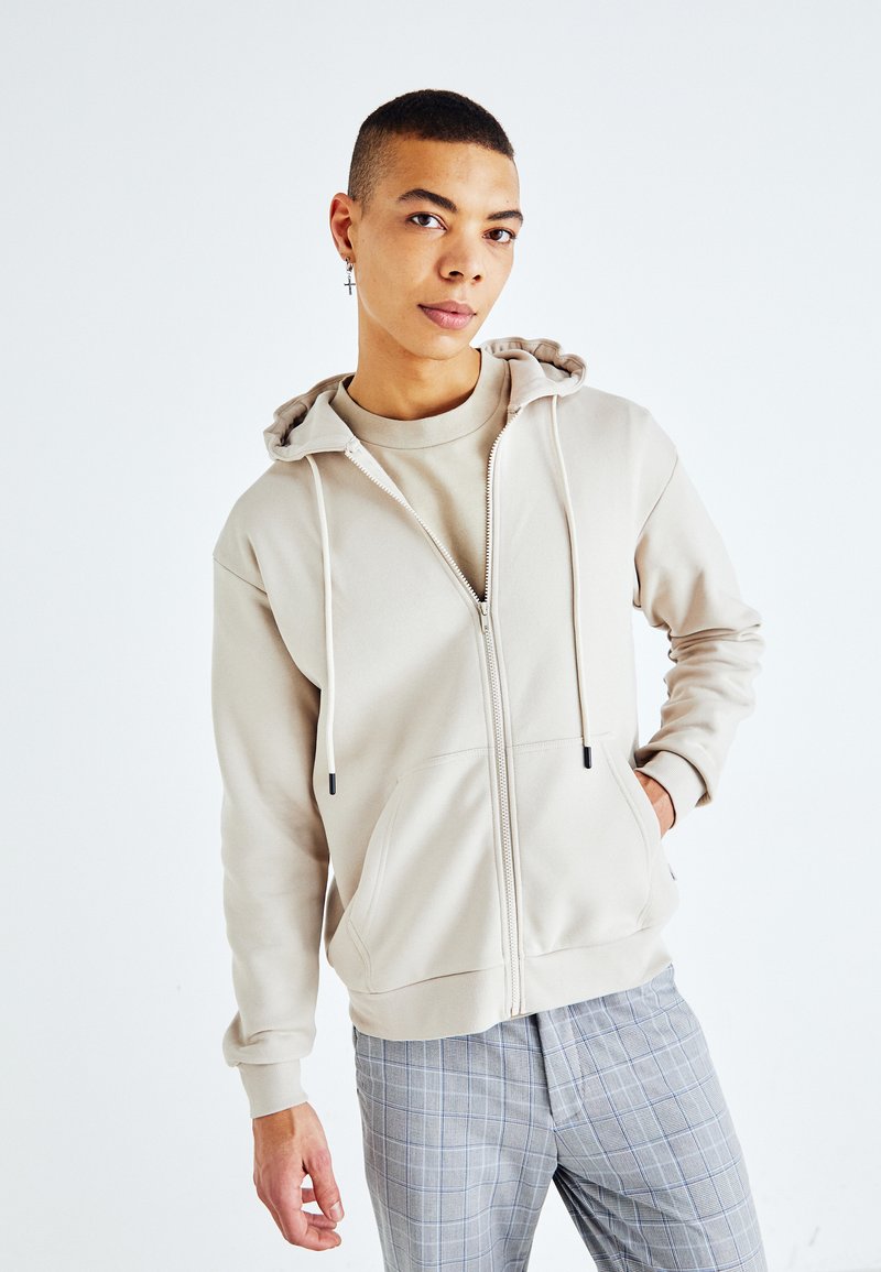 Sudadera beige con cremallera y capucha ajustable, con un diseño liso y bolsillos frontales. Combinada con pantalones de cuadros grises.