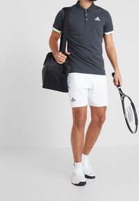 Homme portant un polo Adidas noir et un short blanc, tenant une raquette de tennis noire et un sac à bandoulière, debout devant un fond blanc uni.