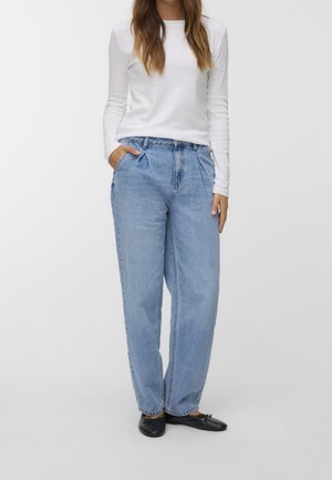 Kvinde iført lyseblå høj talje plisserede jeans, hvid langærmet top og sorte ballerinaer, stående med den ene hånd i lommen.