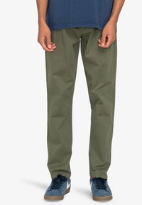 Pantaloni verde oliva con un design a gamba dritta. Realizzati in cotone, dotati di tasche anteriori e una superficie liscia. Indossati con scarpe da ginnastica blu.