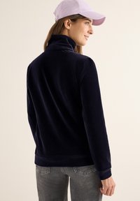 Sudadera de pana azul marino con un cuello alto, mangas largas y un corte relajado. Acompañada de una gorra de béisbol de color púrpura claro.