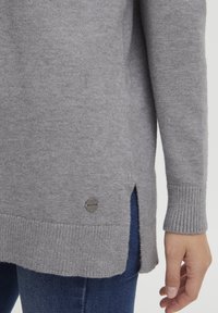 Maglione grigio a maglia con polsini a costine, spacchi laterali e un piccolo logo metallico vicino all'orlo, mostrato con jeans blu.