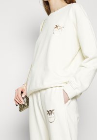 Person wearing cream-colored sweatshirt and matching pants with metallic gold bird logos, holding a small gold chain purse.