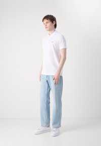 Young man standing, wearing white polo shirt, light blue jeans, and white sneakers, against plain white background.