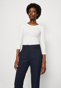 Maglia bianca a maniche lunghe con scollo rotondo, abbinata a pantaloni scuri in tessuto con vita alta e tasche frontali.