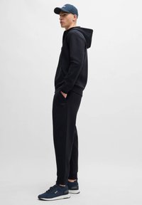 Black hoodie with front pocket, matching black sweatpants, and navy sneakers. The cap is also navy with a logo. Casual athletic wear.