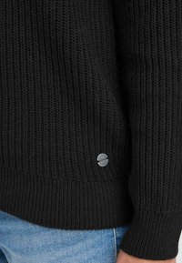 Pull en tricot noir avec des poignets et un ourlet côtelés, arborant une petite étiquette logo en argent sur le côté. Tissu texturé visible. Denim visible en dessous.