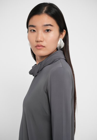 Grå langærmet bluse med drapering ved kraven, glat overflade og tætsiddende silhuet. Modellen bærer store sølvøreringe.