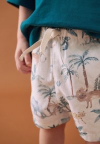 Shorts en tissu léger avec un imprimé animalier tropical représentant des lions et des palmiers. Taille élastique avec un cordon crème. Chemise bleu sarcelle visible.