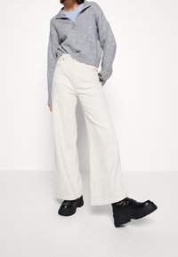 Kremowe spodnie w szerokim kroju z teksturowanym wykończeniem, w połączeniu z jasnoszarym swetrem zapinanym na zamek i masywnymi czarnymi butami na platformie.