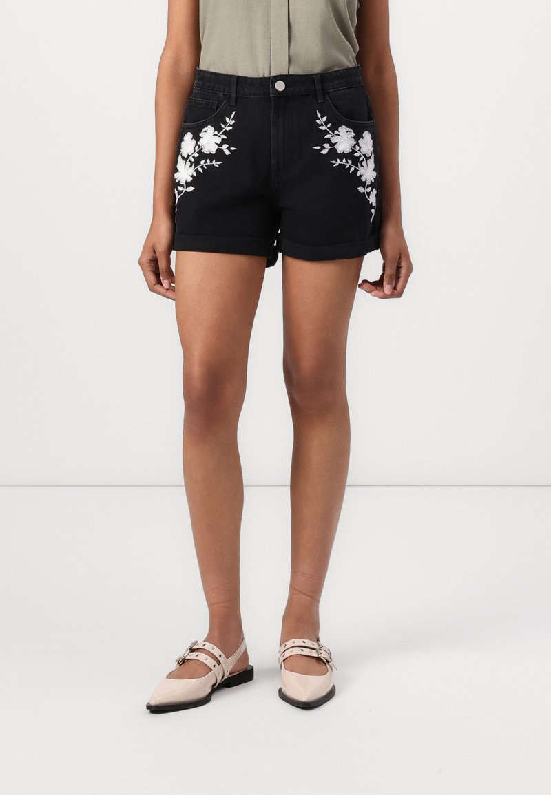 Svarte denimshorts med blomsterbroderi på lommene, oppbrettede kanter og frontknapplukking. Kombinert med lysebeige flate sko.