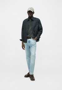 Must ruuduline särkkleit tumeda halli T-särgi kohal, helesinised teksad, pruunid nahast kingad ja beež nokamüts.