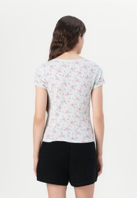 Camicetta a maniche corte di un leggero colore menta con motivo floreale rosa e righe verticali. Presenta un colletto rotondo e un tessuto a trama. Indossata con pantaloni corti neri.