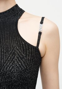 Κοντινό πλάνο ενός ώμου που φοράει ένα μαύρο γκλίτερ ριμπ halter τοπ με λεπτή ρίγα που φέρει μεταλλικό λογότυπο "Versace Jeans Couture".