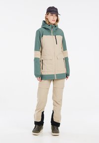Beige und grüne Skijacke mit Kapuze, die über zwei Fronttaschen und einen Reißverschluss verfügt. Beigefarbene Skihose mit Cargo-Taschen und schwarzen Bündchen.
