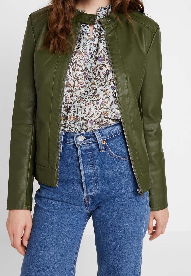 Giacca in pelle verde con zip, dal design aderente, abbinata a una blusa floreale e jeans di denim blu a vita alta.