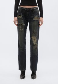 Blugi albăstriți închis cu aspect uzat, detalii rupte și accente metalice cu ținte pe buzunarele din față. Materialul are o textură ușor uzată.