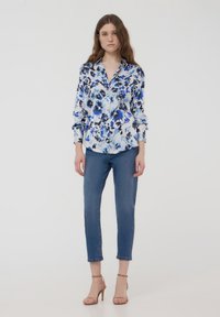 Camicia colletto in pattern floreale blu su sfondo bianco, abbinata a jeans slim-fit blu e sandali con tacco beige.