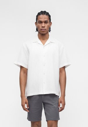 Young man with braided hair wearing a white short-sleeve button-up shirt and gray knee-length shorts, standing against a plain background.