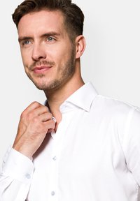 Biała koszula z teksturowanej tkaniny, z wąskim kołnierzykiem i niebieskimi guzikami, noszona przez mężczyznę, który poprawia kołnierzyk, prezentująca widok z bliska.