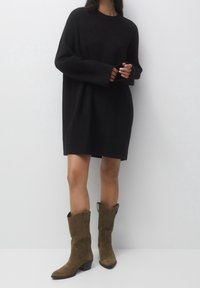 Robe pull noire oversize avec manches longues, en tissu texturé et coupe décontractée, associée à des bottes de cow-boy en daim vert olive.