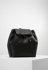Mochila de cuero negro con una superficie texturizada, cierre de cordón y una solapa adornada con tachuelas. Presenta acentos de hardware plateado.