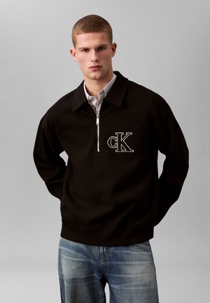 Jeune homme portant un sweat-shirt noir à fermeture éclair courte avec un logo "cK" blanc sur la poitrine et un jean bleu délavé, debout devant un fond gris.