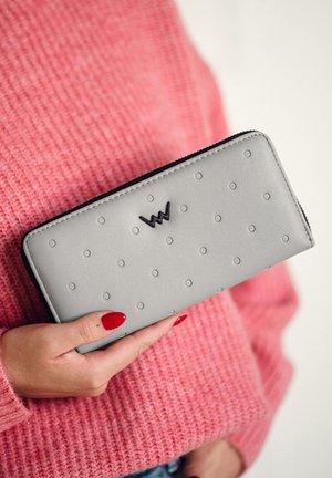 Gray textured wallet with raised dot pattern, zip closure, and black logo. Held in hand, with a pink knit sweater in the background.
