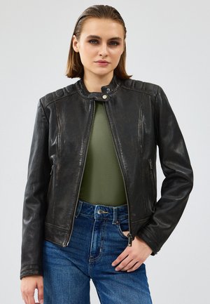 Zwarte leren jas met een staande kraag, gewatteerde schouderaccenten, ritssluiting aan de voorkant en zijzakken. Draag een groene top eronder.