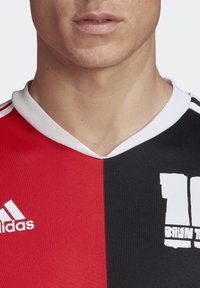 Jersey deportivo rojo y negro con cuello en V, collar blanco, que presenta un logo blanco y un diseño de números en negrita en la sección negra.