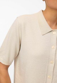 Ljust beige kortärmad stickad polo med en texturerad yta, med en spetsig krage och fem neutrala knappar framtill.