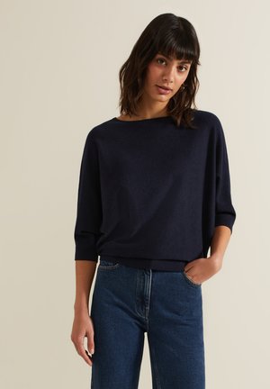 Vrouw met donker haar die een losse marineblauwe trui met driekwartmouwen en blauwe spijkerbroek draagt, staat met één hand in de zak.