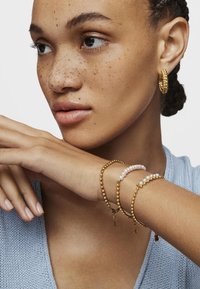 Pendientes de aro de oro y tres pulseras: cuentas de oro, perlas de agua dulce, y cuentas alternando oro y blanco. El fondo es neutral y claro.
