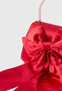 Punainen satiininen käsilaukku, jossa on ylisuuri rusetti ja valkoisista helmistä valmistettu kahva, yksivärisellä taustalla.