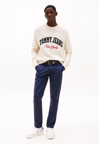 Kremgenser med "TOMMY JEANS New York" i blått og rødt, kombinert med marineblå bukser og hvite joggesko. Avslappet passform. Ensfarget bakgrunn.