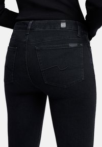 Schwarze Jeans aus Denim mit tailliertem Design, ausgestattet mit einem Logo-Patch auf der Rückseite, auffälliger Taschennähte und glatter Textur.