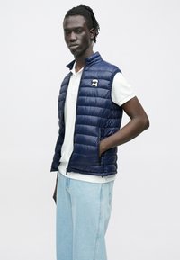 Mann iført en marineblå vattert vest over en hvit polotrøye og lyseblå jeans, stående med en hånd i vestlommen mot en ensfarget bakgrunn.