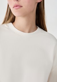 Sudadera de color beige claro con cuello redondo, hecha de una tela suave con una textura lisa y detalles acanalados en el cuello.