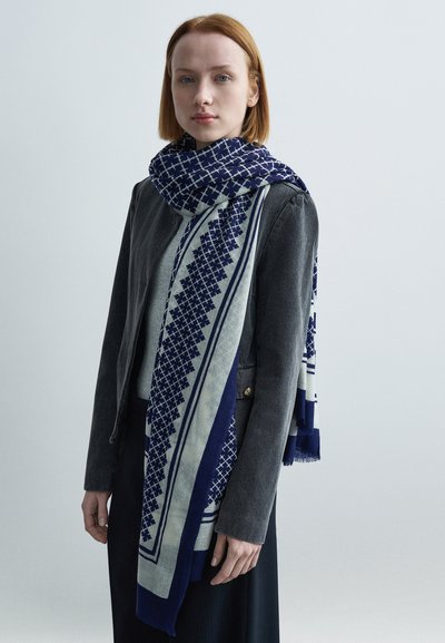 Écharpe à motifs bleu marine et crème avec des dessins géométriques, en tissu texturé, drapée sur une veste grise, avec des bords frangés.