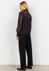 Kvinna som står i profil klädd i en mörklila långärmad spetsblus, svarta vida byxor och svarta ballerinaskor mot en enfärgad bakgrund.