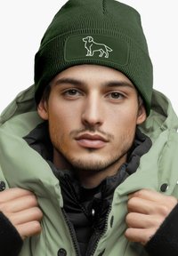 Green knitted beanie with a textured finish and a dog silhouette patch. Worn with a light green puffy jacket, layered over a black top.