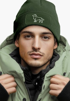 Green knitted beanie with a textured finish and a dog silhouette patch. Worn with a light green puffy jacket, layered over a black top.