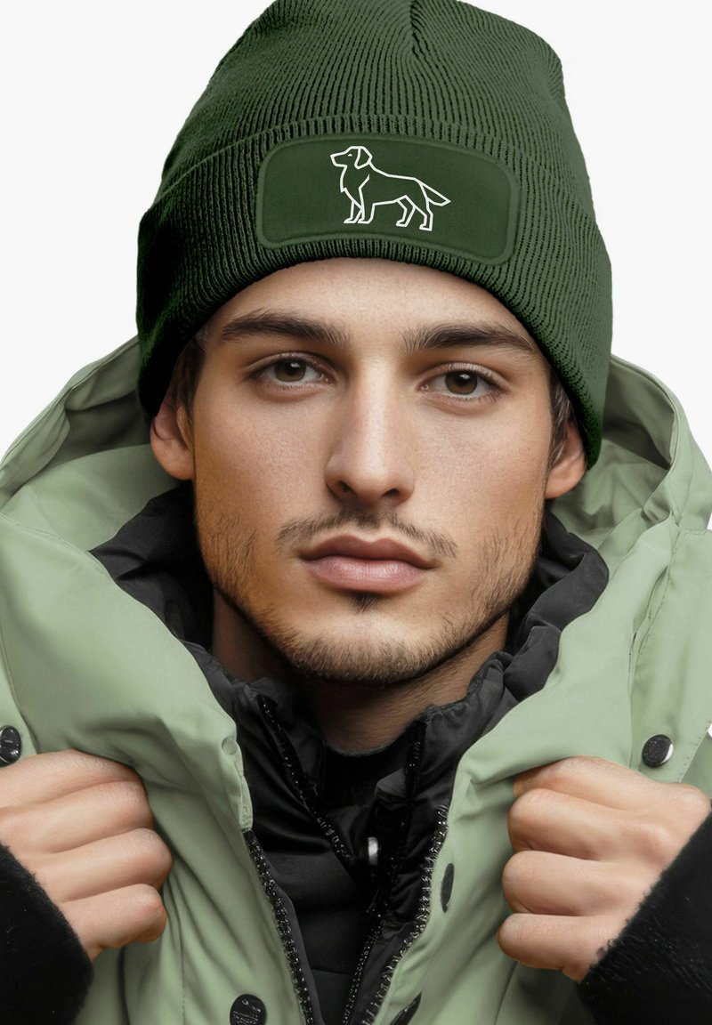 Green knitted beanie with a textured finish and a dog silhouette patch. Worn with a light green puffy jacket, layered over a black top.
