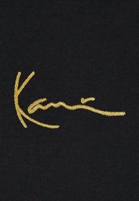 Black fabric displaying a gold embroidered signature design, featuring smooth, bold lines and a slightly raised texture.