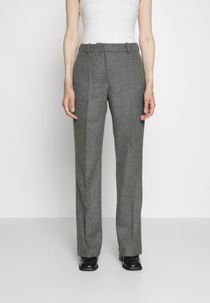 Persona que lleva pantalones grises de tiro alto y pierna recta con pliegues delanteros, zapatos negros y una camiseta blanca sin mangas, de pie sobre un fondo liso.