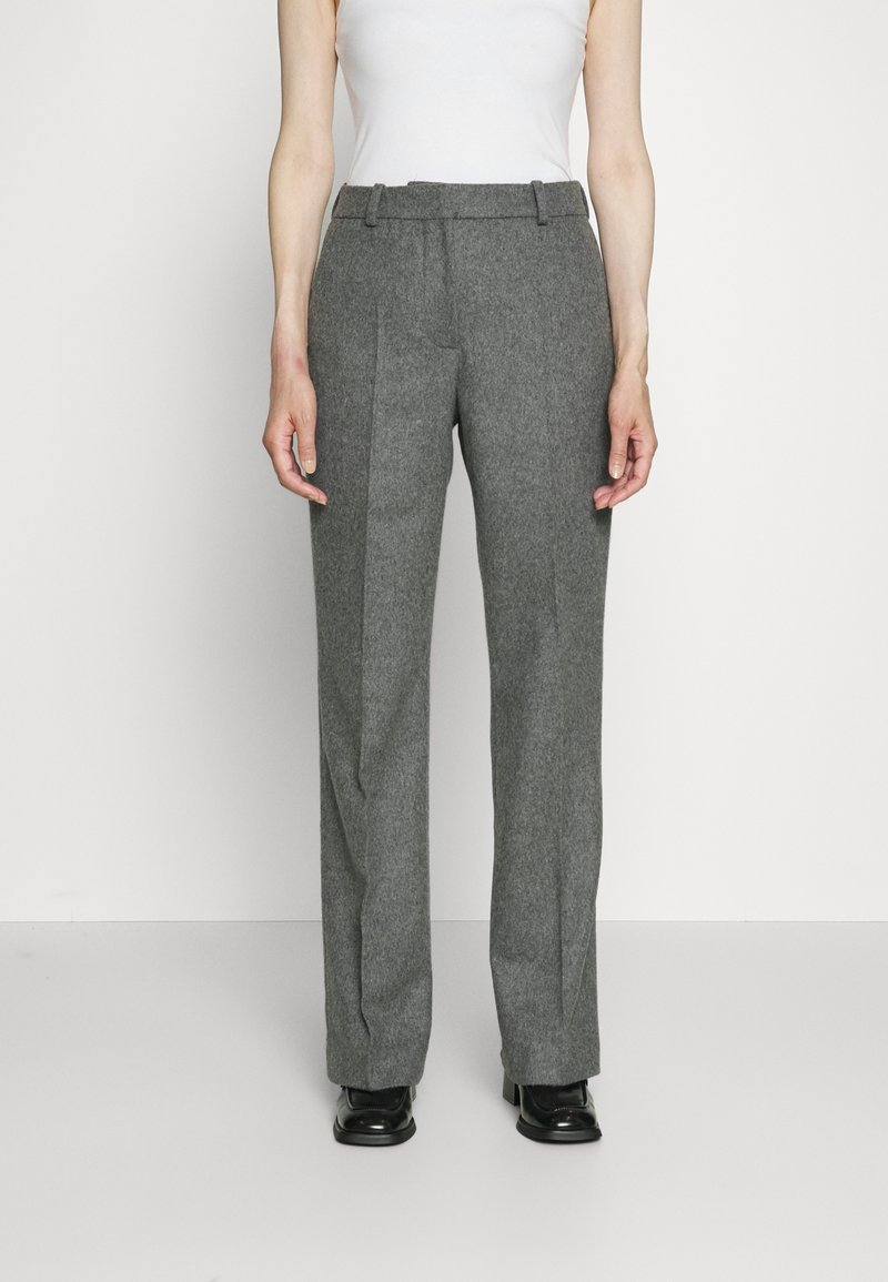 Personne portant un pantalon gris taille haute à jambes droites avec des plis sur le devant, des chaussures noires et un haut blanc sans manches, debout sur un fond uni.