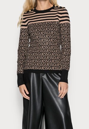 Gestreifter Strickpullover in Schwarz und Beige mit "GUESS"-Muster, goldenen Knopf-Details an den Schultern und gerippten Bündchen. Kombiniert mit schwarzen Hosen.